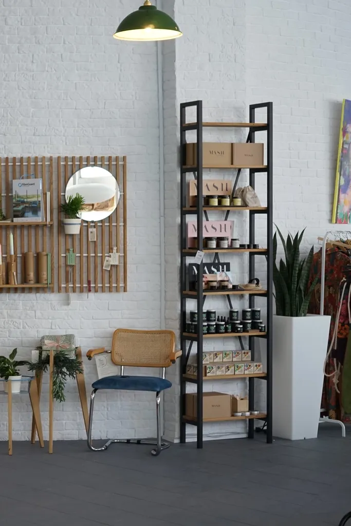 Stylowe wnętrze z czarną metalową półką z produktami do pielęgnacji skóry, drewnianym krzesłem z wiklinowym siedziskiem, wysoką rośliną w białej doniczce i dekoracyjnymi elementami ściennymi, w tym okrągłym lustrem i oprawionym obrazem. Jasny i nowoczesny wystrój z wiszącą lampą.